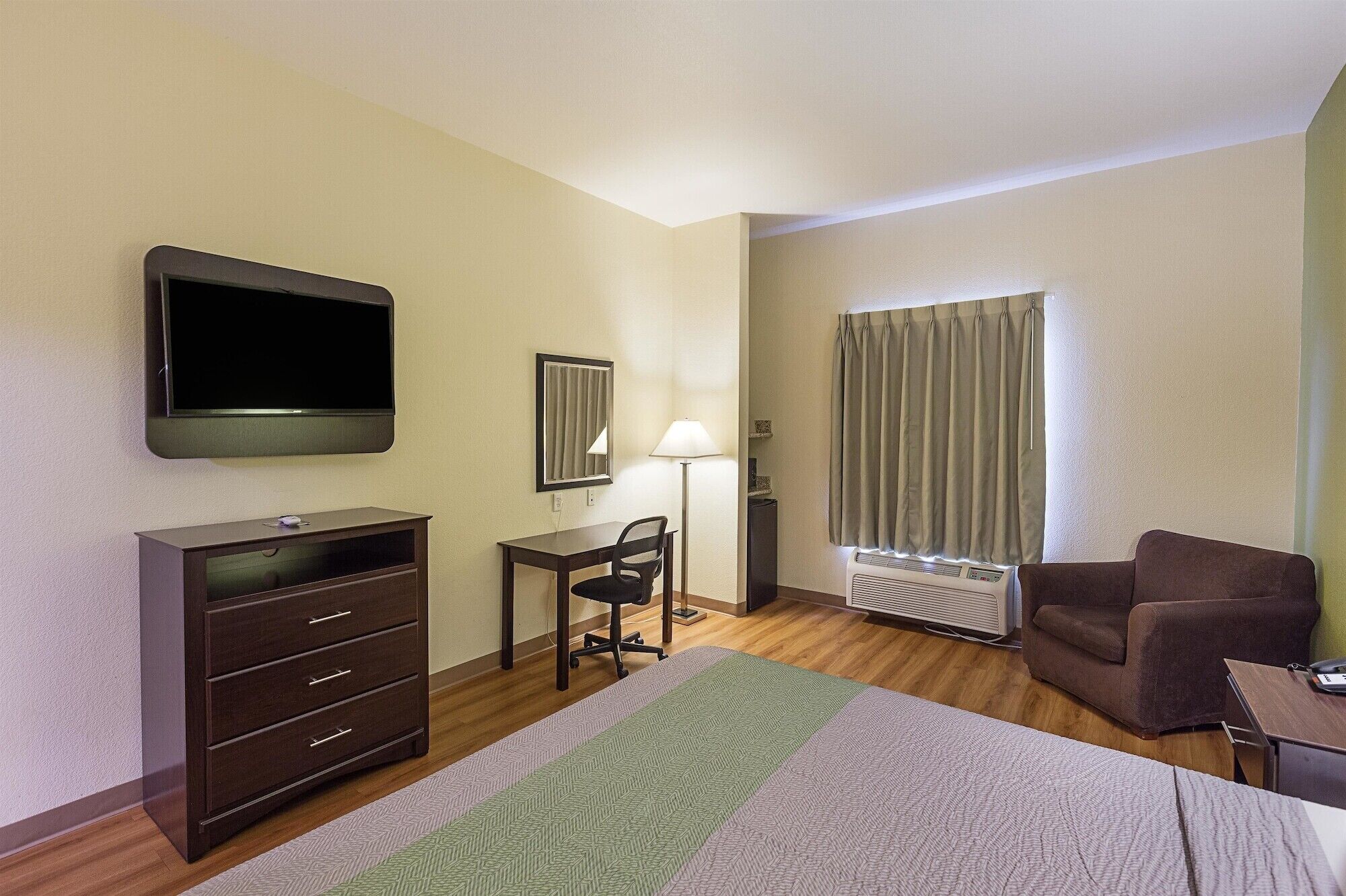 A hotel room with a bed, a desk with a chair, a dresser, a television, a window with curtains, and a radiator.