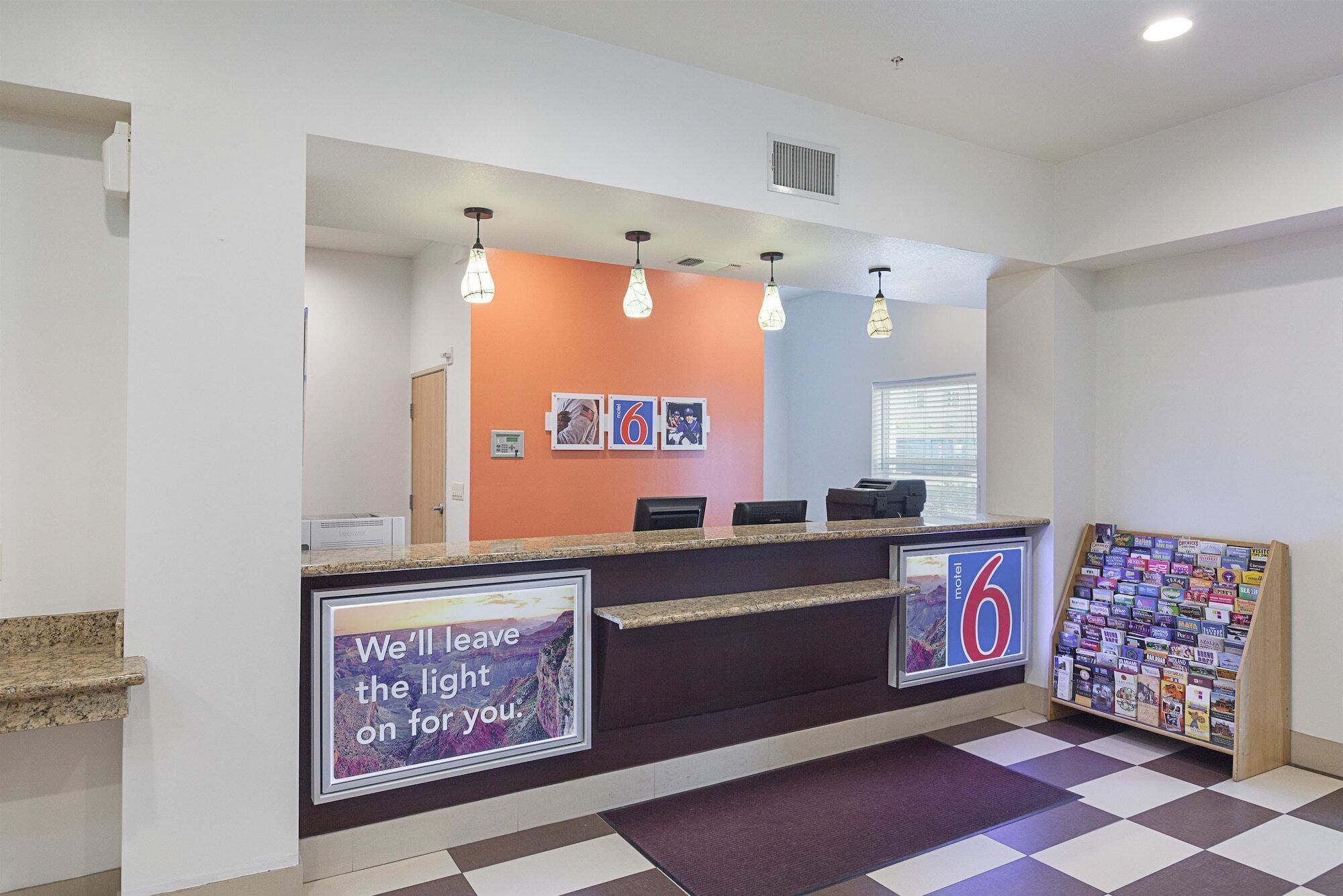 Uma área de recepção de hotel com balcão de check-in, um mural de avisos com folhetos de viagem e uma parede com fotografias emolduradas e uma placa.