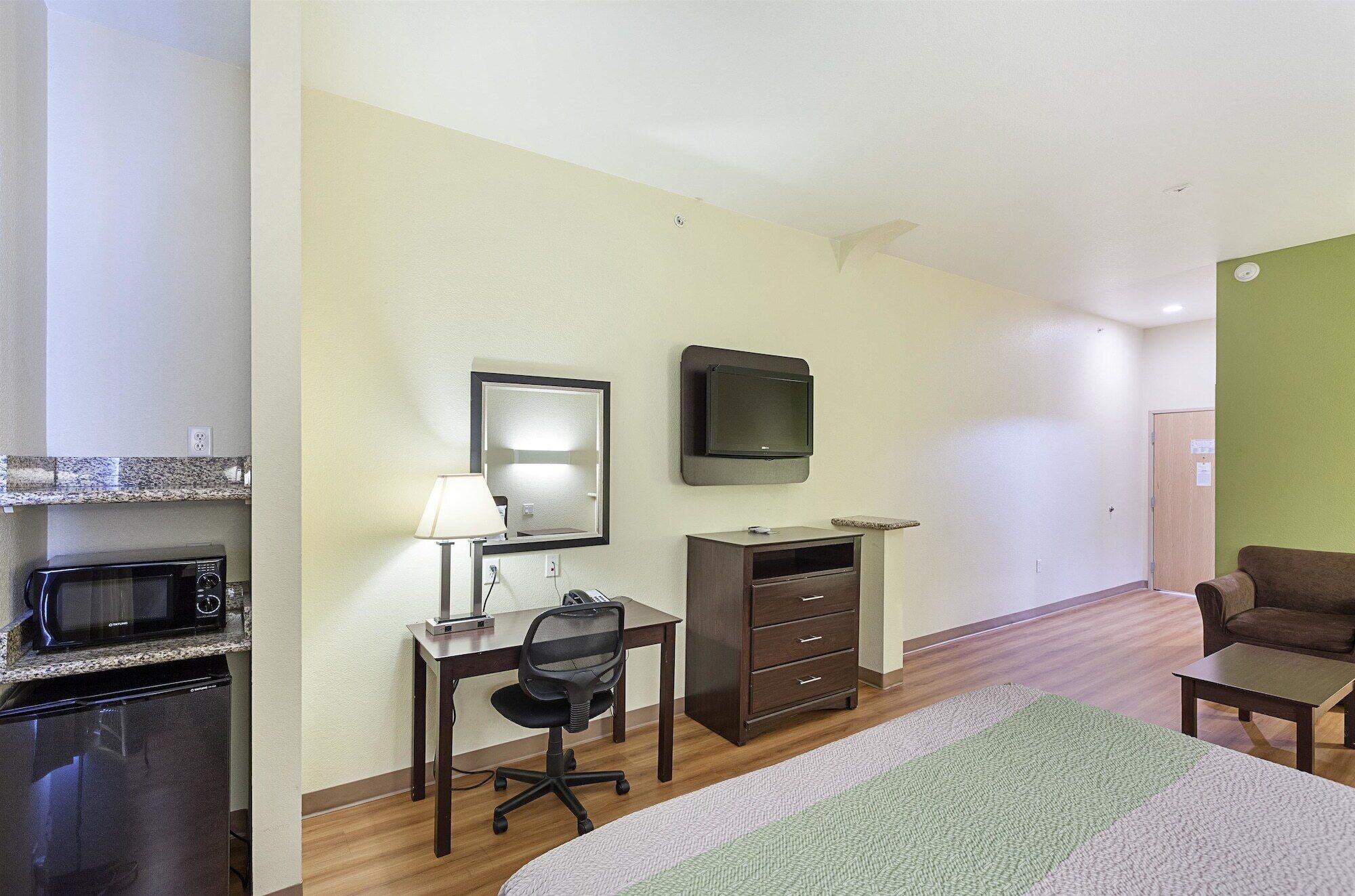 A hotel room with a kitchenette, a desk with a chair, a television, a dresser, and a bed.