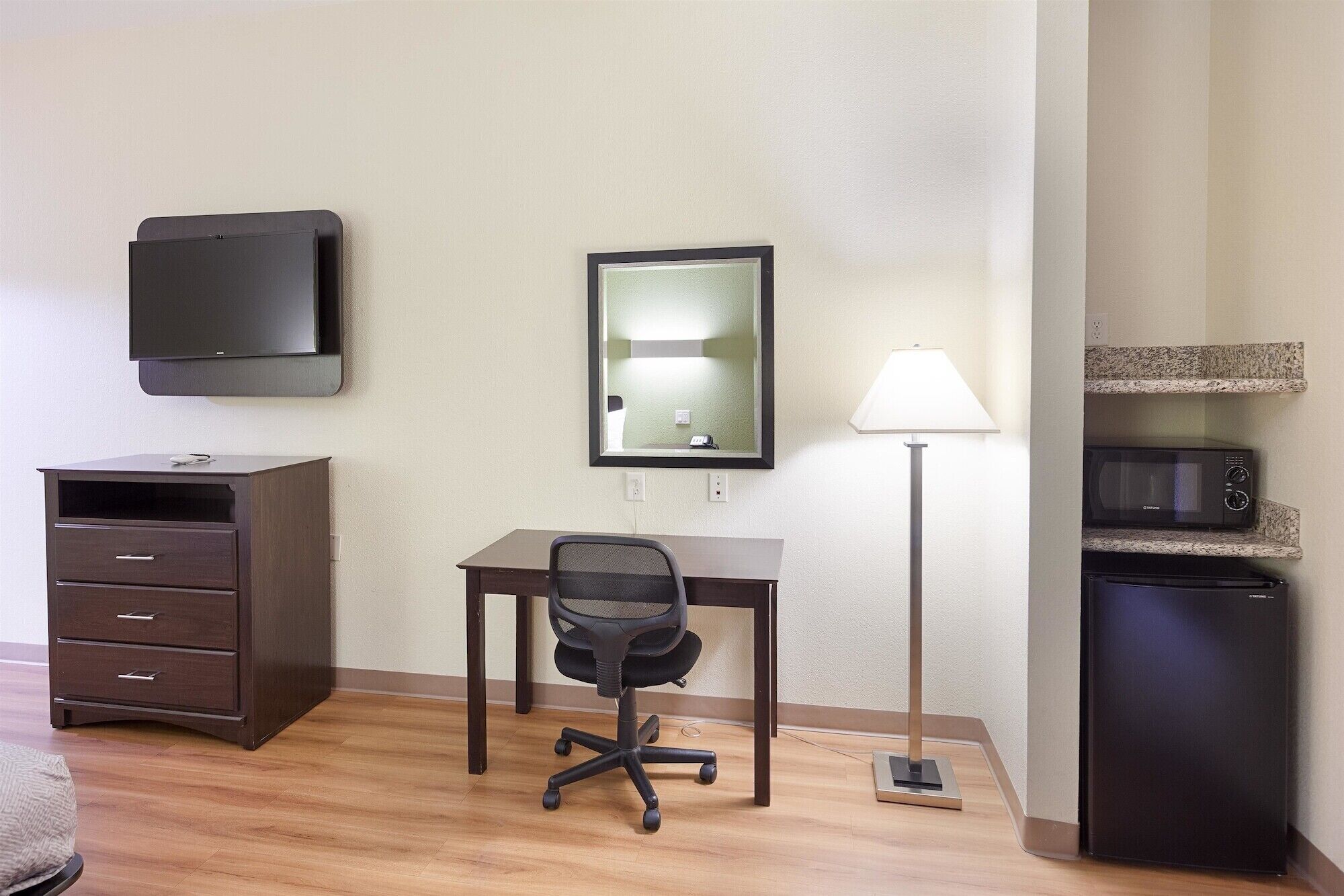 Habitación de hotel con escritorio, silla, televisor, espejo, lámpara y kitchenette.