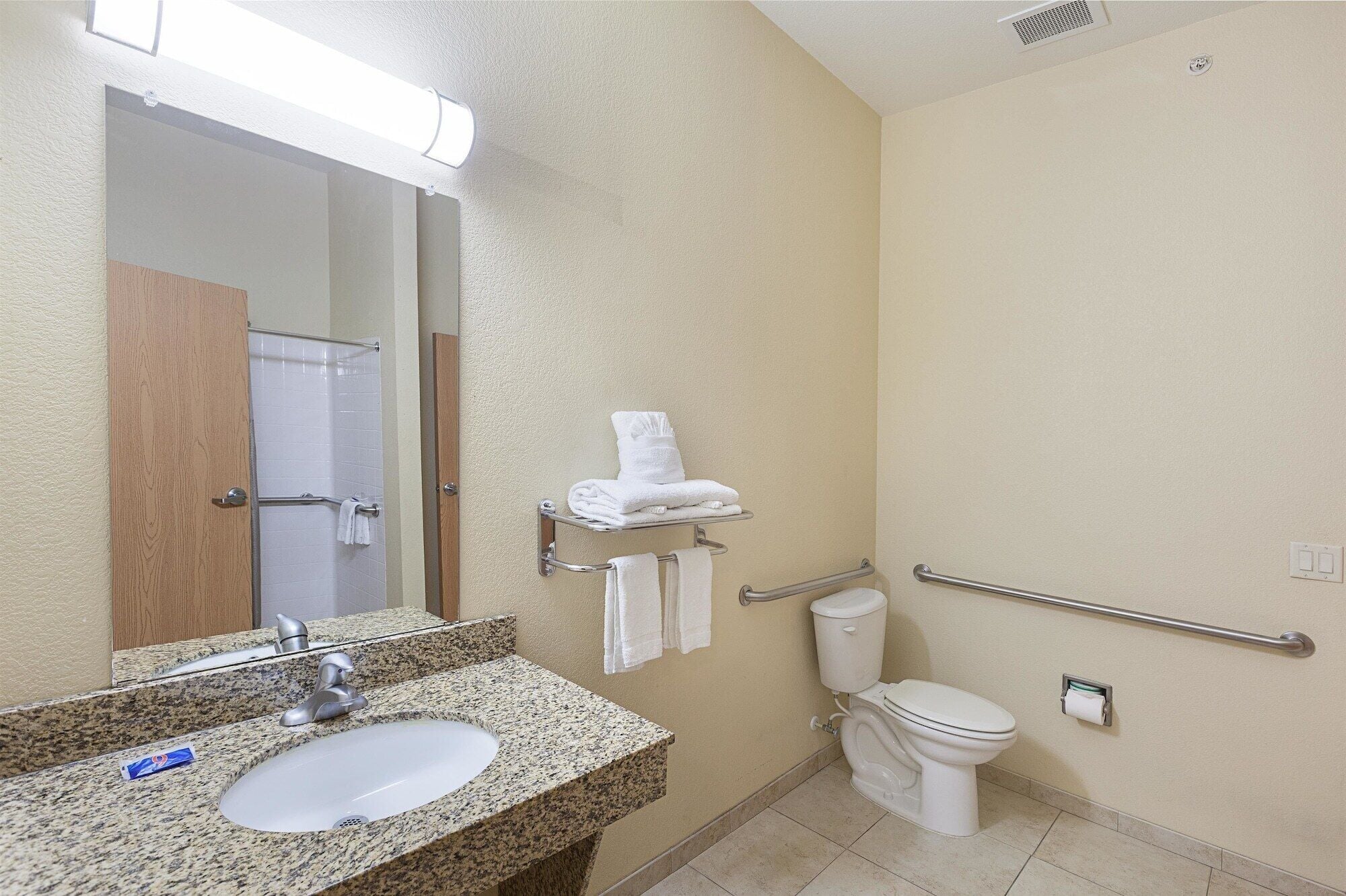 A bathroom with a granite countertop sink, a mirror, a towel rack, a toilet, and a handrail.