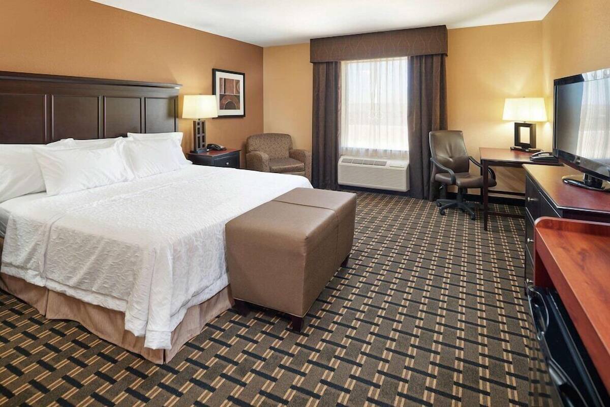 A hotel room with a large bed, a desk with a television, a chair, an air conditioning unit, and a patterned carpet.