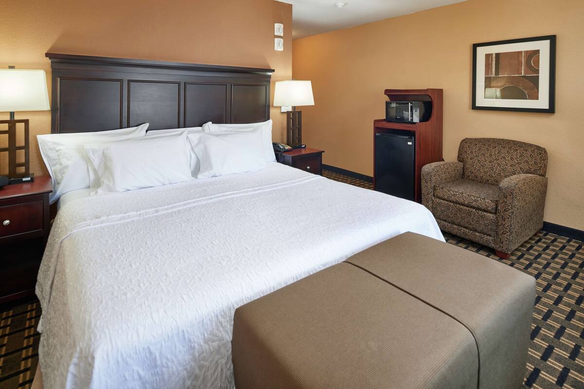 A hotel room with a large bed, a chair, a desk, a lamp, and a microwave.