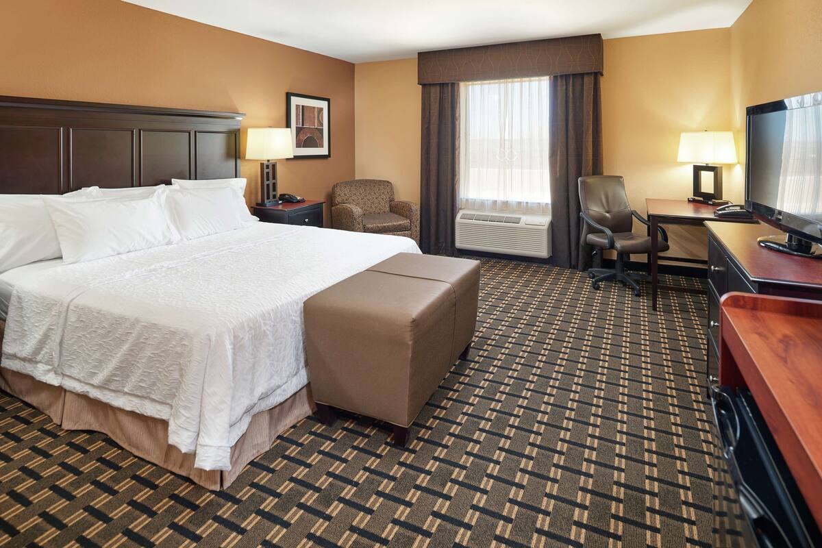 A hotel room with a large bed, a desk with a television, a chair, and an air conditioning unit.
