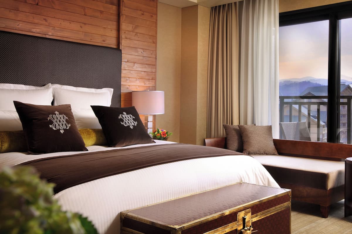 Habitación de hotel con una cama grande, una cama más pequeña, cabecera de madera, mesita de noche, lámpara, ventana con vista a montañas y un jarrón decorativo con flores.