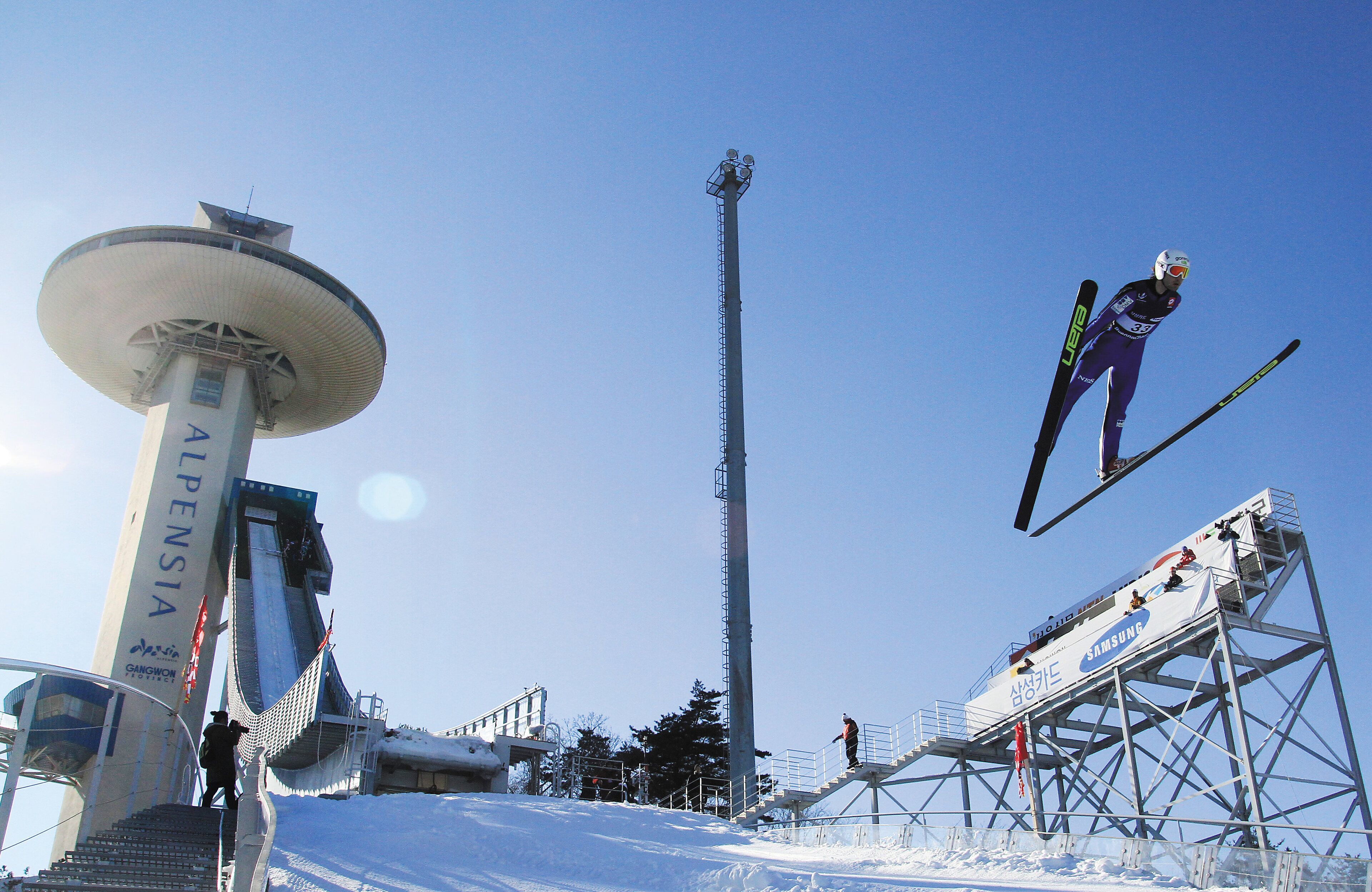 Una rampa de salto de esquí con un anuncio de Samsung y una alta torre con la leyenda 'ALPENSIA' al fondo.