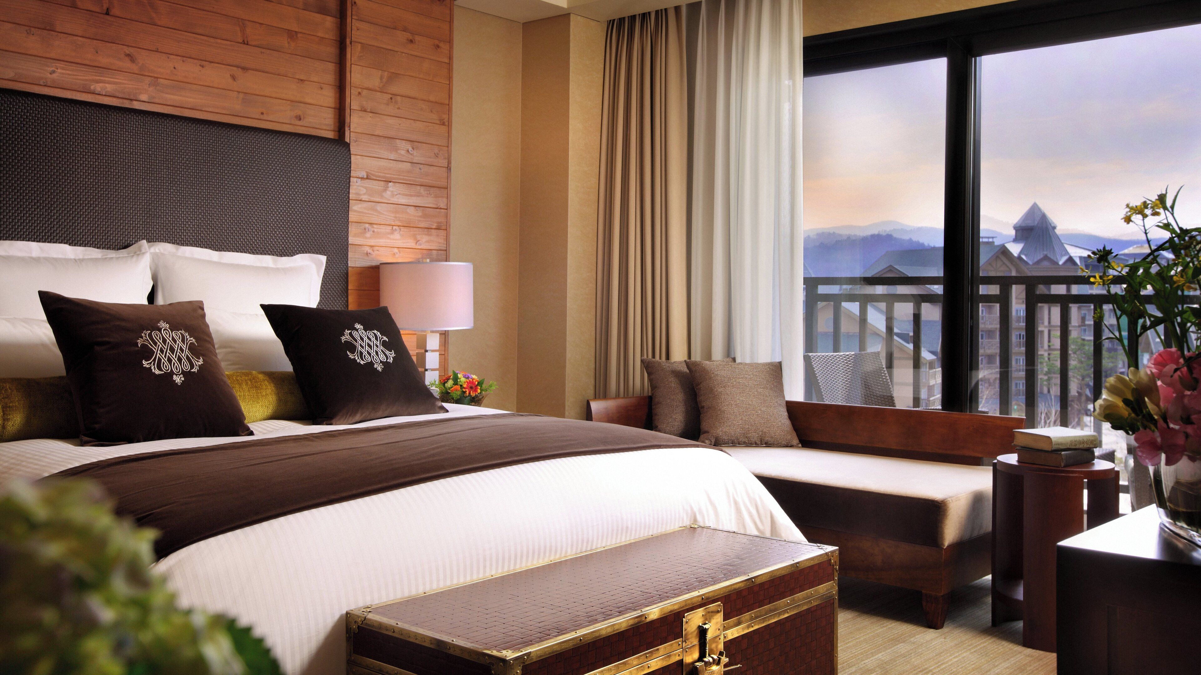 Hotelkamer met een groot bed, een kleiner bed, een houten hoofdeinde, een nachtkastje, een lamp, een raam met uitzicht op bergen, en een decoratieve vaas met bloemen.