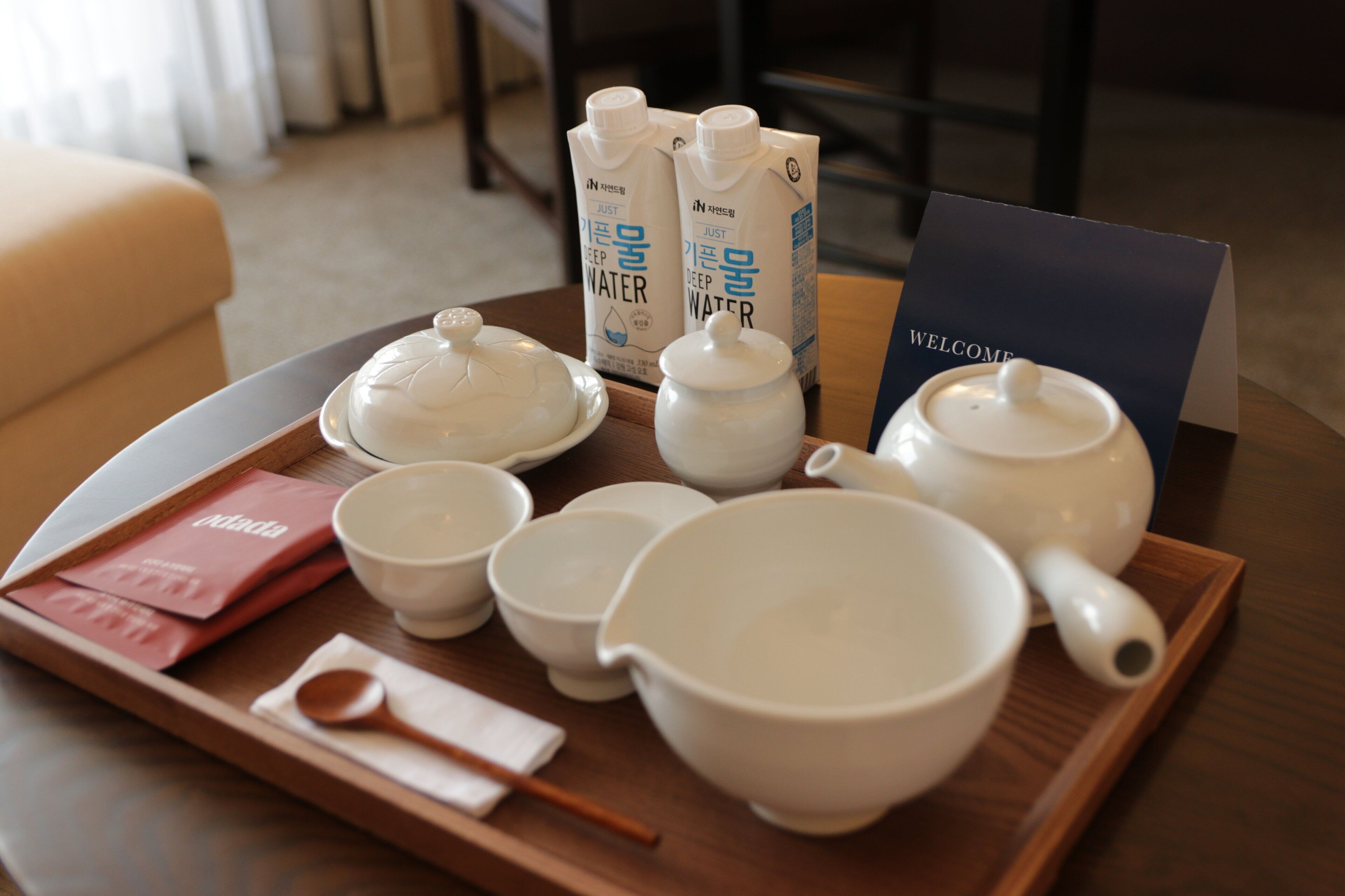 A tray with tea set and water bottles on a table, a welcome card, and a red item.