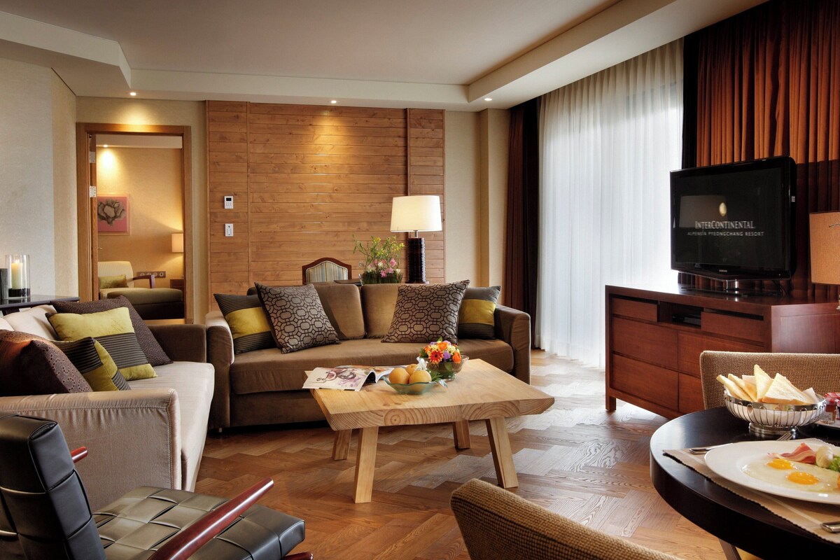 A modern hotel room with a sofa, a dining table with breakfast, a TV, and a wooden wall.