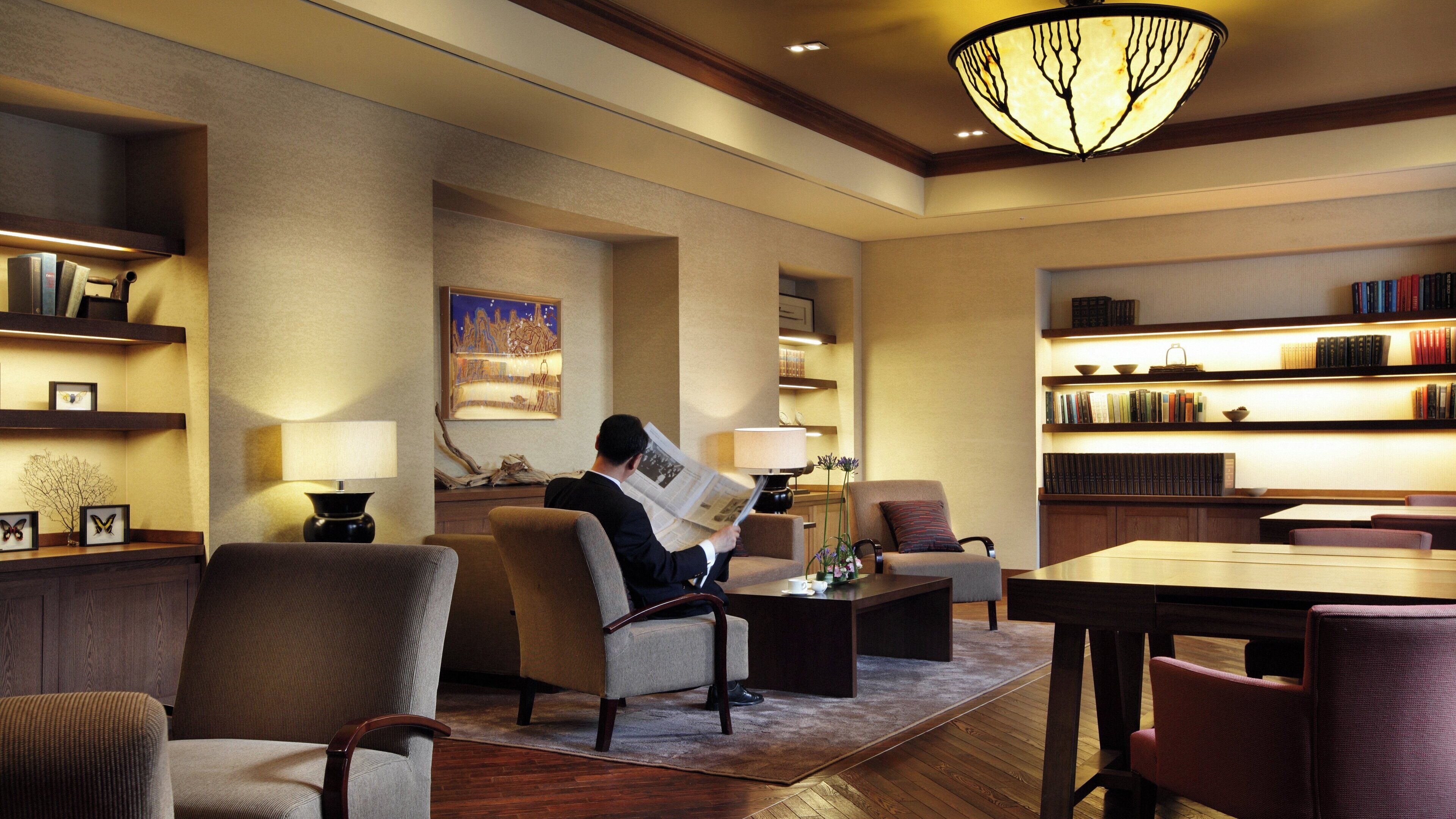A hotel lobby with a man reading a newspaper, a fireplace, bookshelves, and a chandelier.