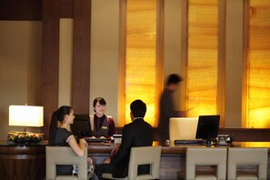 A hotel reception desk with a receptionist, a guest, and a man in a suit. There is a computer, a telephone, and a small table with a lamp and a bowl of fruit.