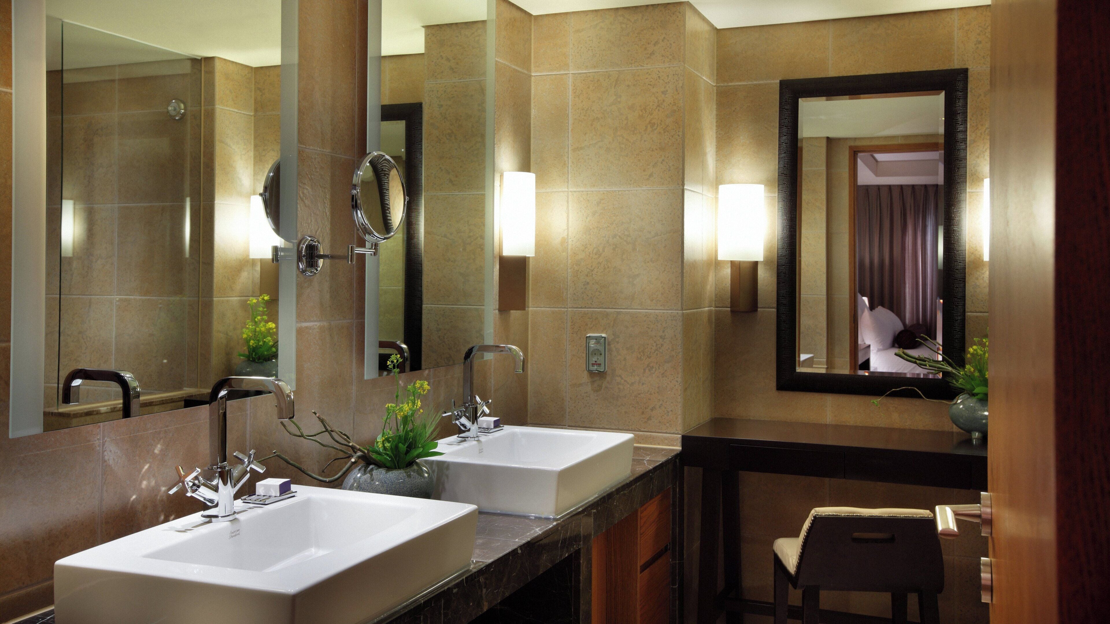 A hotel bathroom with two sinks, a mirror, and a chair.