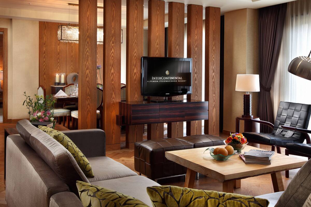 Una sala de estar moderna con un sofá, una mesa de centro con un libro y flores, un televisor sobre un soporte de madera, y un área de comedor al fondo.