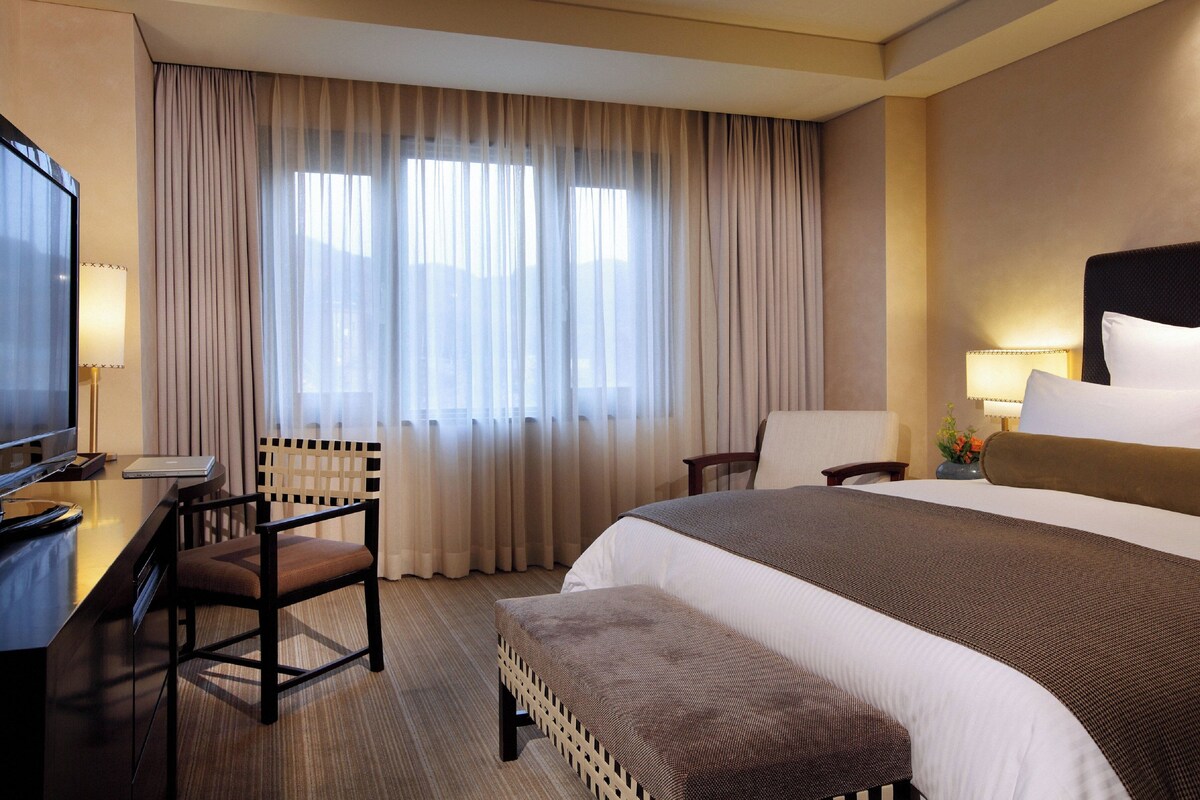 A hotel room with a bed, a desk with a chair, a television, and a window with curtains.