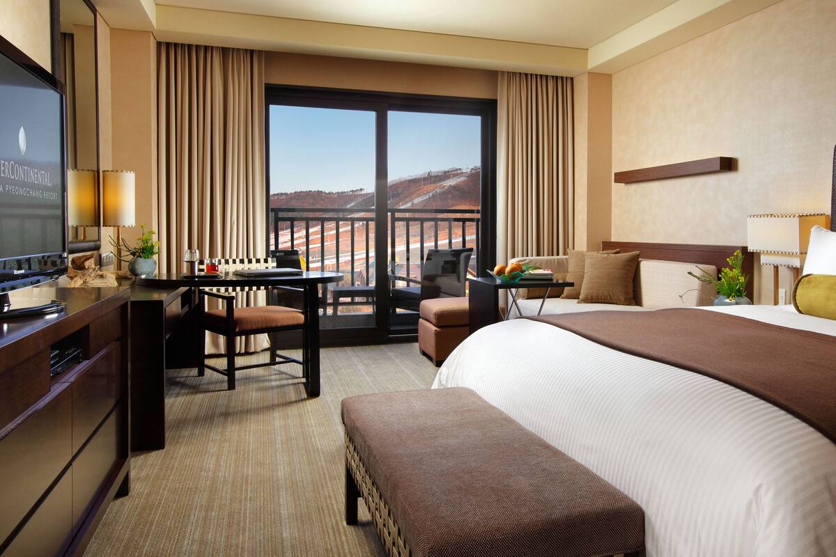 Habitación de hotel con una cama grande, un escritorio con computadora, una silla, un televisor y vista a un paisaje montañoso.