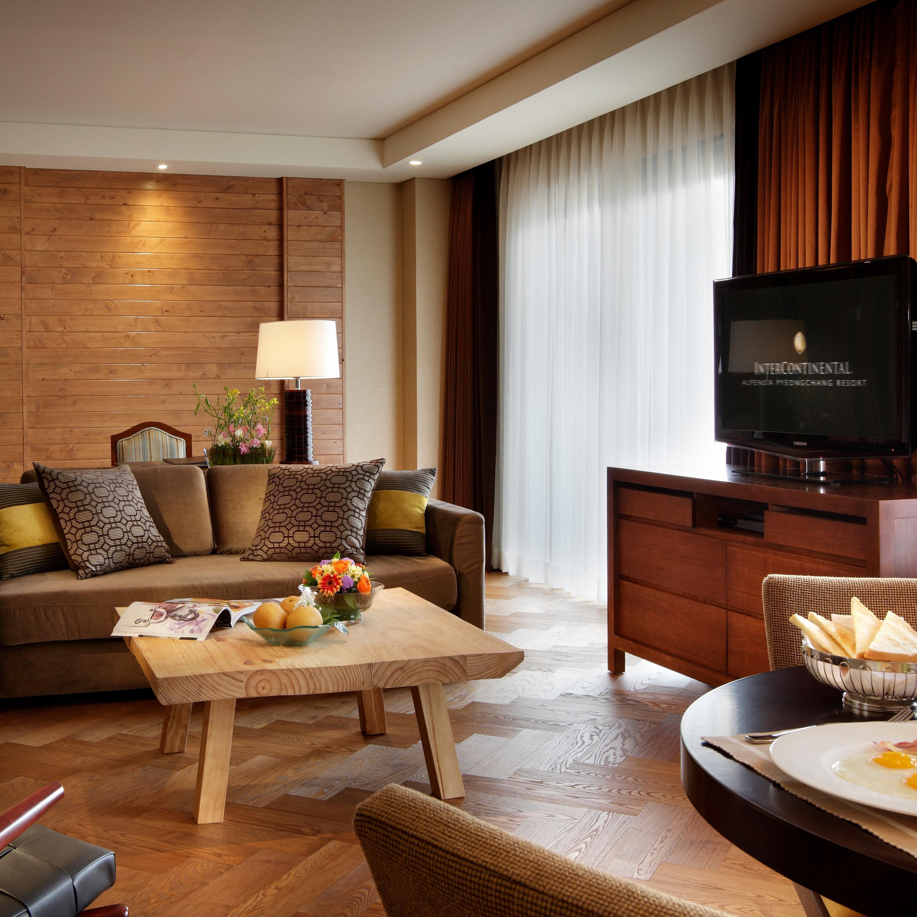 A modern hotel room with a wooden panel wall, a sofa with patterned cushions, a coffee table with a flower vase, a dining area with a plate of food, and a television displaying "INTERCONTINENTAL".