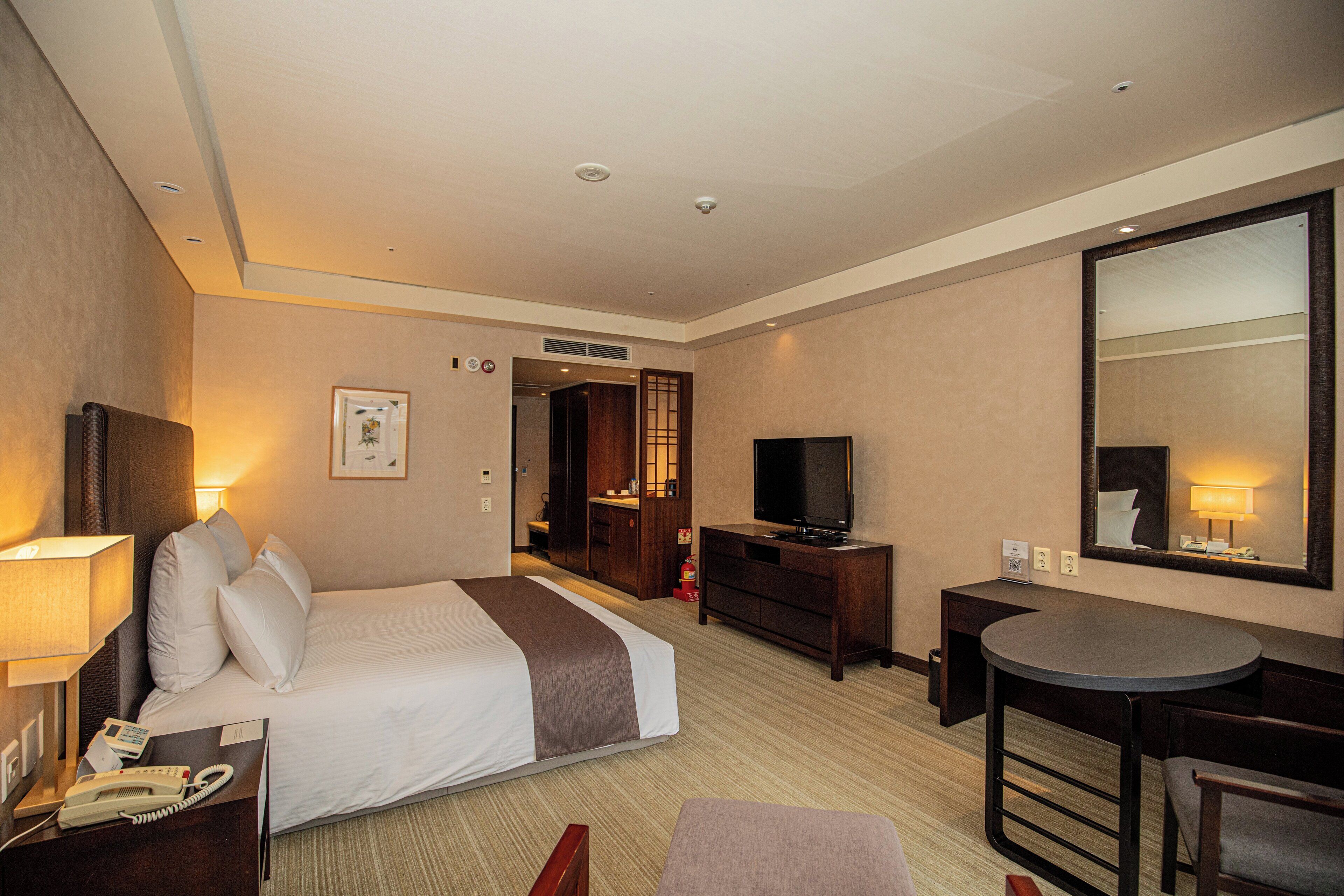 A hotel room with a bed, bedside table with a lamp, a TV, a desk with a chair, and a mirror.