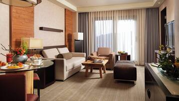 Habitación de hotel con sofá, sillón, otoman, mesa de centro y zona de comedor con una mesa preparada para el desayuno.