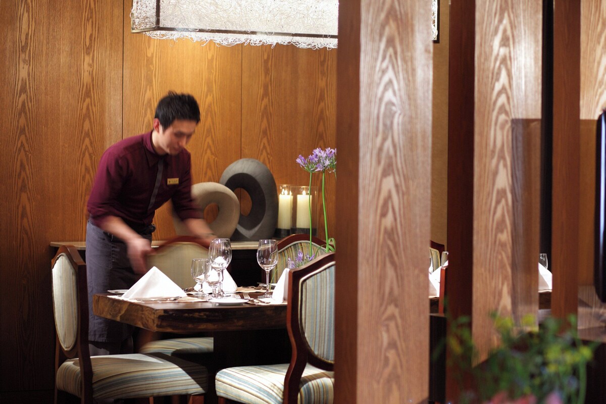 A dining area with a wooden table set for two, a television on a stand, and a candle on the table.