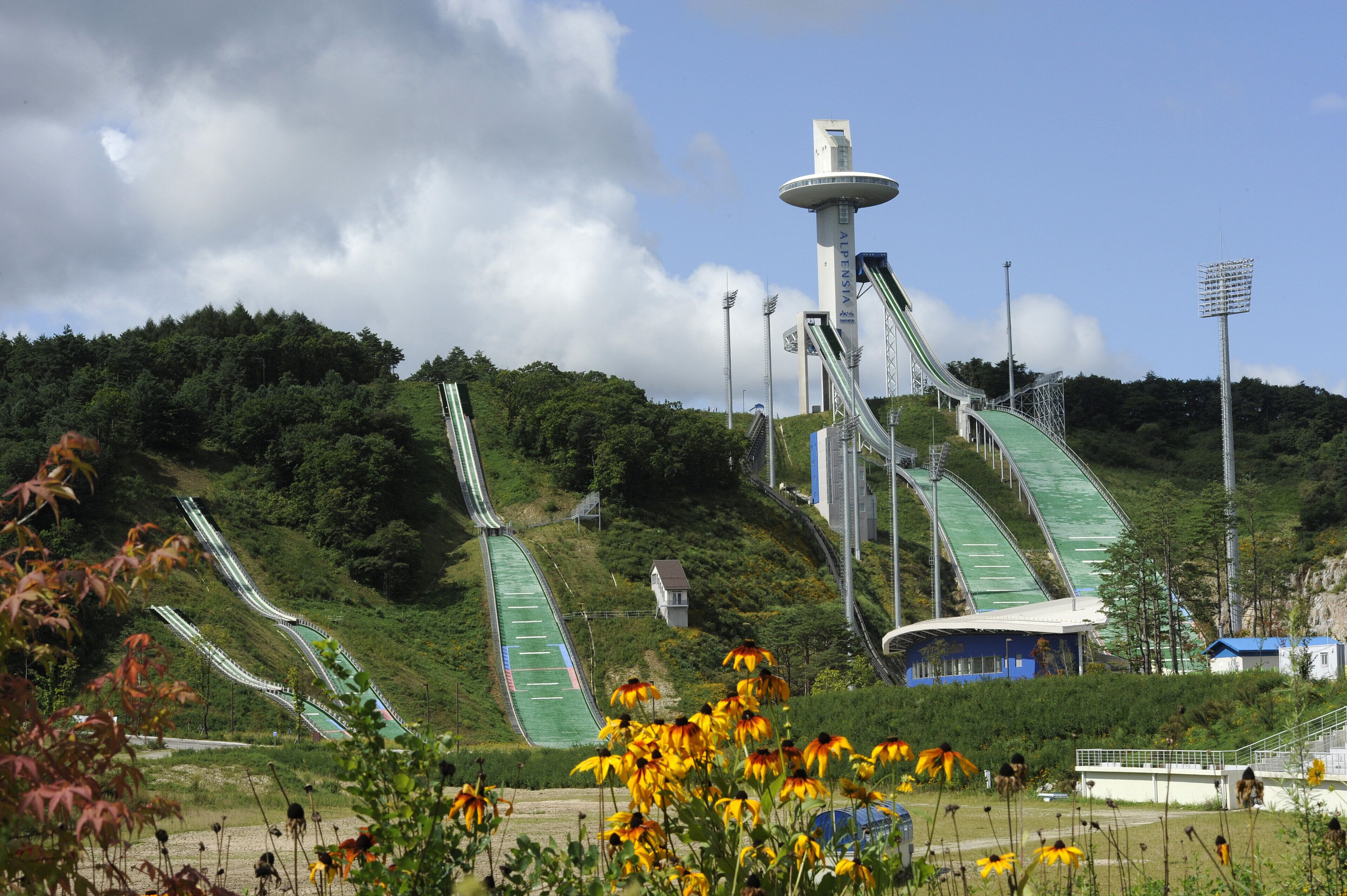 A ski lift system with multiple chairlifts and a central tower, surrounded by greenery and a clear sky.