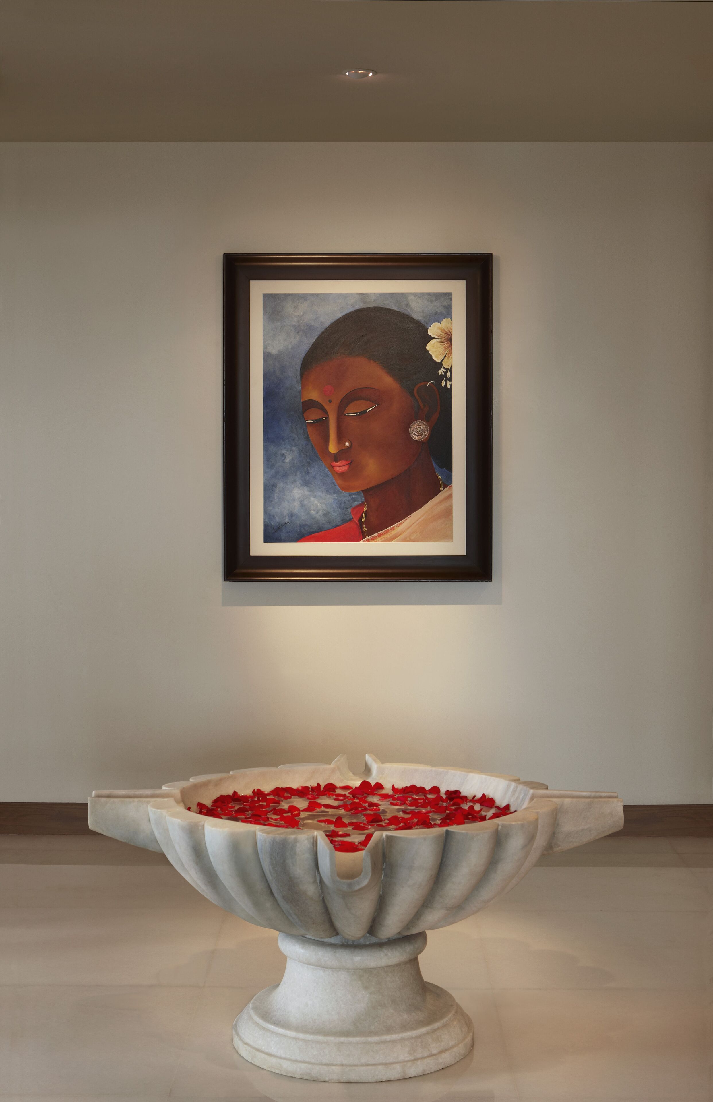 Una obra de arte en marco sobre una pared, encima de un cuenco de piedra lleno de flores rojas.