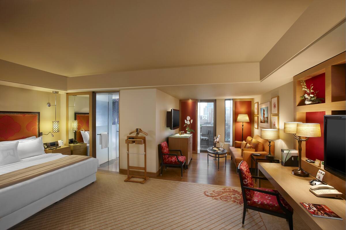 A hotel room with a bed, a desk, a chair, a sofa, a TV, and a bathroom with a mirror and towel rack.