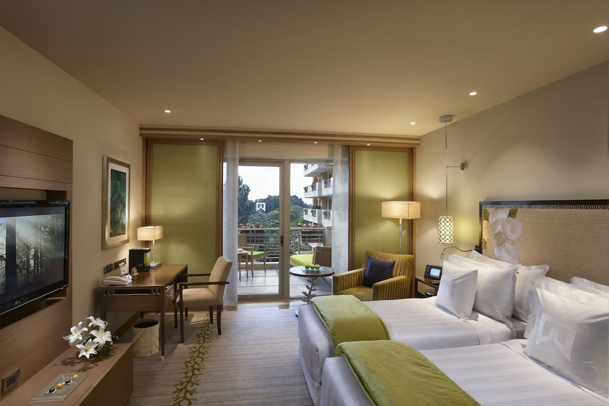 A hotel room with a bed, a desk with a computer, a TV, and a balcony with a view of buildings.