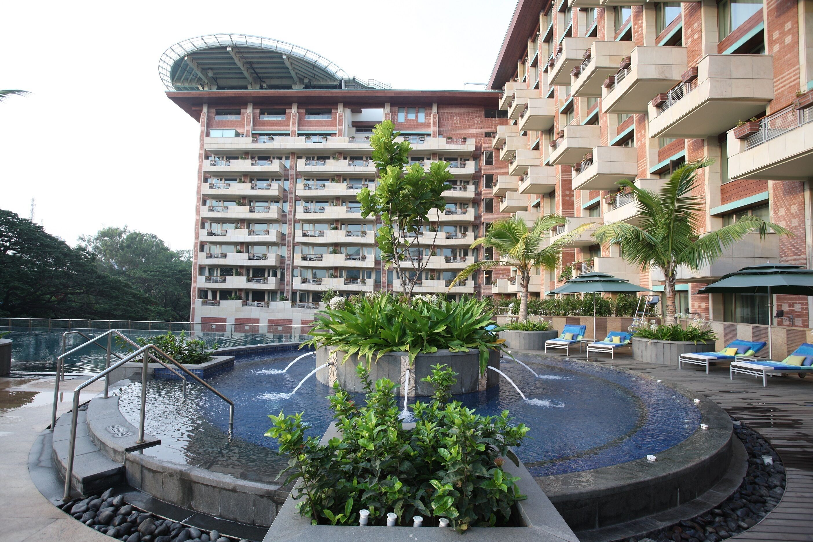 Zona de piscina de hotel con una fuente circular, sillones de descanso y un edificio grande con varios balcones al fondo.