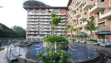 Zona de piscina de hotel con una fuente circular, sillones de descanso y un edificio grande con varios balcones al fondo.