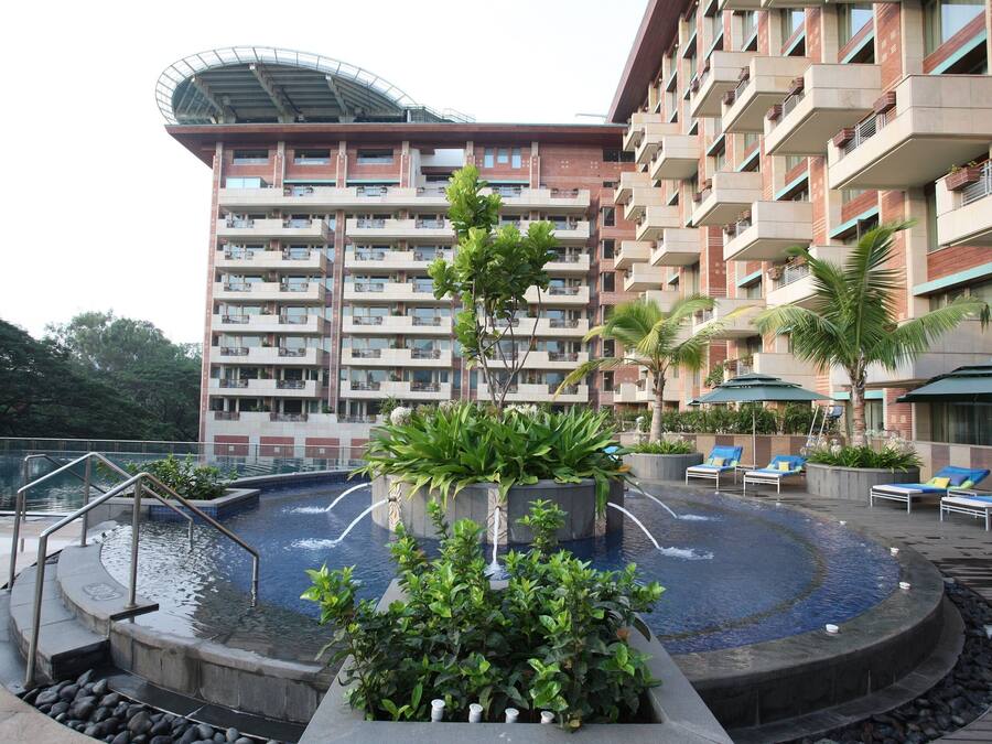 Zona de piscina de hotel con una fuente circular, sillones de descanso y un edificio grande con varios balcones al fondo.