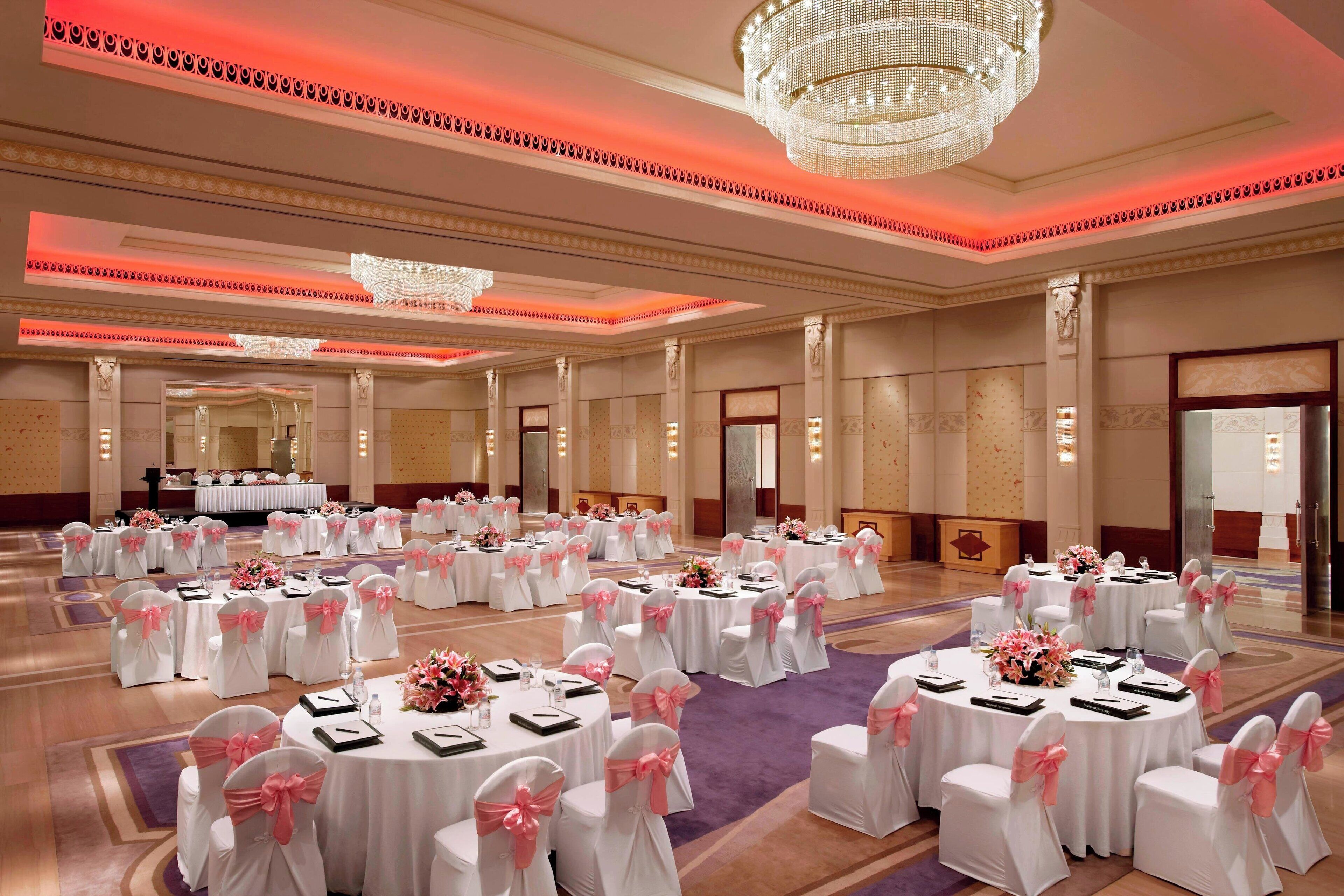 A large banquet hall with round tables, white chairs with pink sashes, and a chandelier.