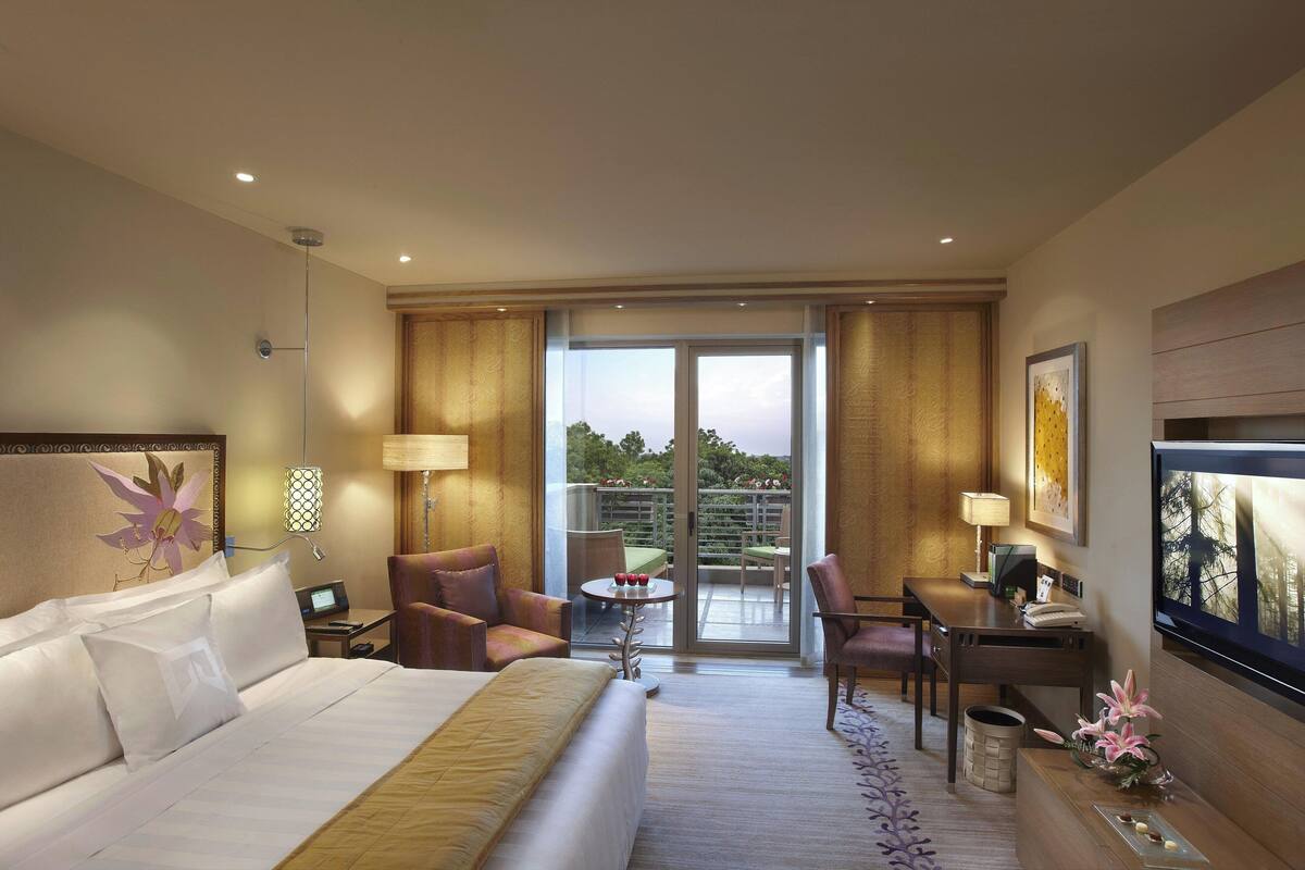 A hotel room with a bed, a desk, a chair, a small table with a lamp, and a TV. There is a balcony with a view of the sky and trees.