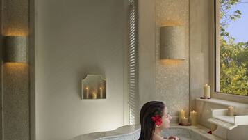 Um quarto de spa com uma banheira grande cheia de água e pétalas de rosa, uma janela com vista para árvores e velas fixadas na parede.