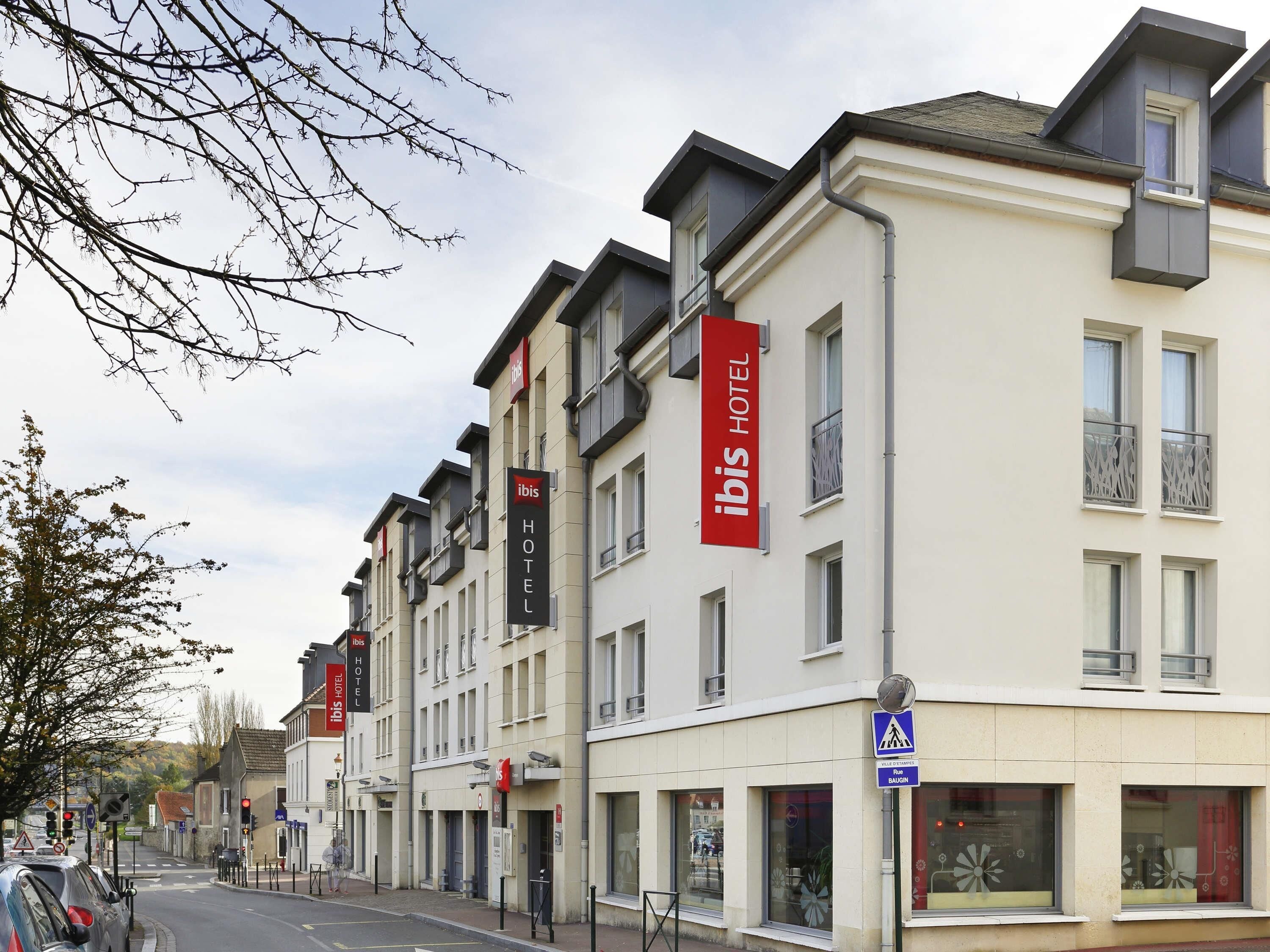 Una fila di edifici moderni di un hotel con la segnaletica 'ibis HOTEL', un attraversamento pedonale e un cartello blu che indica una località nelle vicinanze.