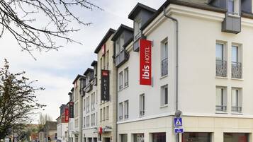 A row of modern hotel buildings with "ibis HOTEL" signage, a pedestrian crossing, and a blue sign indicating a nearby location.