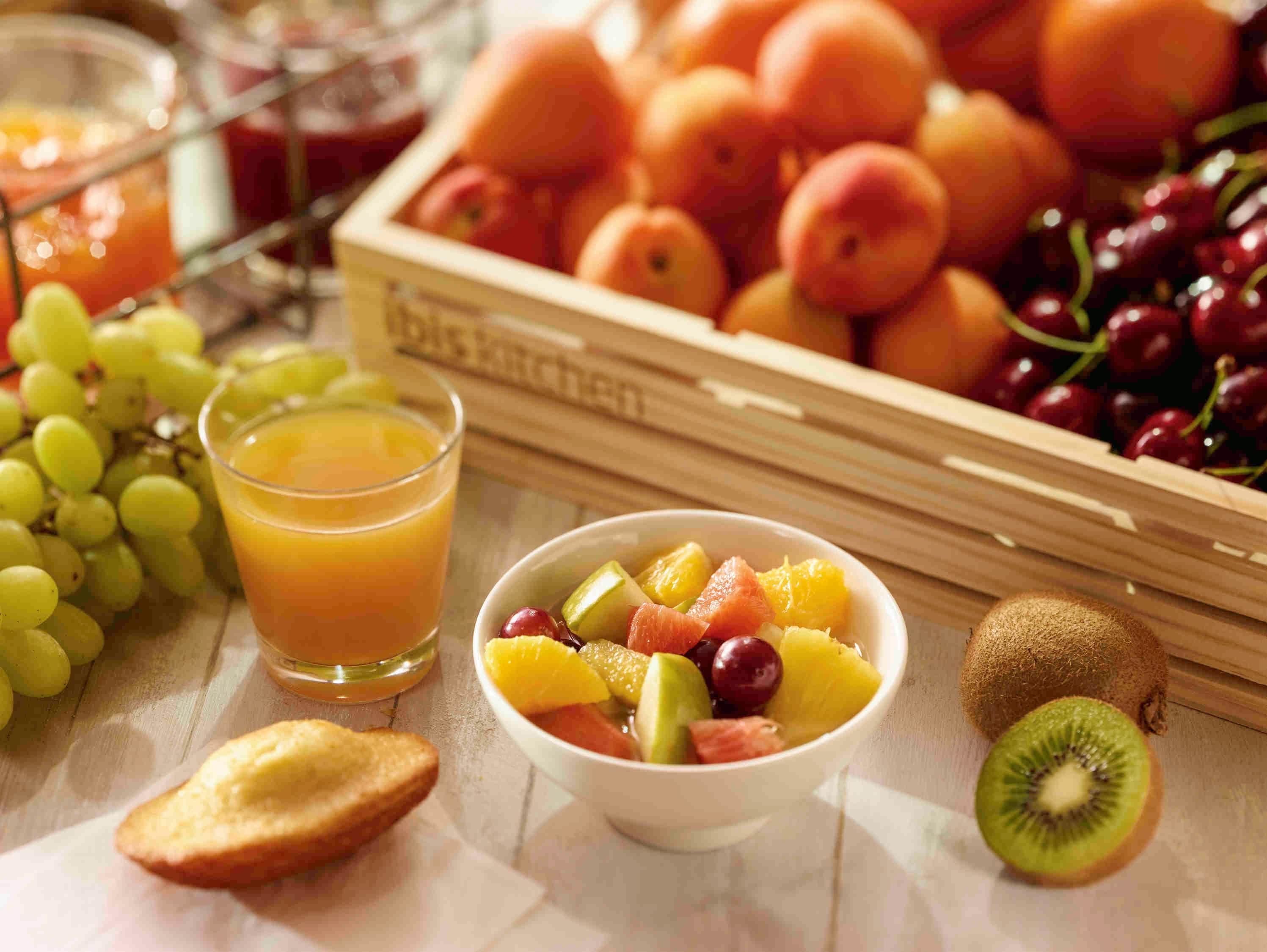 Un cuenco de frutas con uvas, kiwi y otras frutas, un vaso de jugo de naranja y una caja de melocotones.