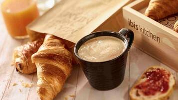 A wooden tray with croissants, a black mug with a creamy beverage, and a glass of orange juice on a wooden surface.