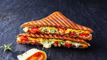 A grilled sandwich with visible ingredients like tomatoes, cheese, and eggs, placed on a dark surface.