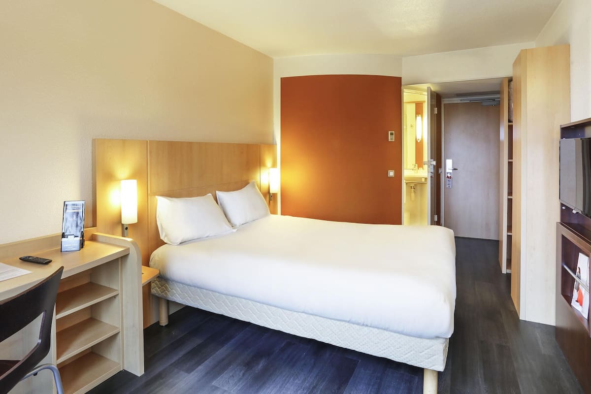 A hotel room with a bed, bedside table, lamp, and a desk with a chair.