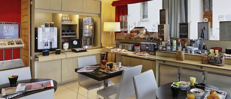 A hotel breakfast area with a coffee machine, breakfast bar, seating, and a view of the street.
