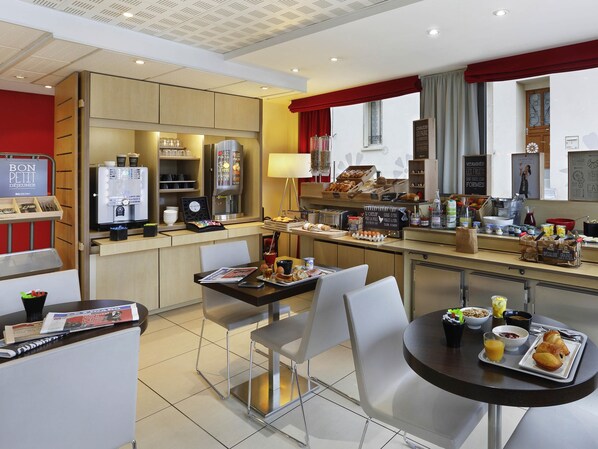 A hotel breakfast area with a coffee machine, breakfast bar, seating, and a view of the street.