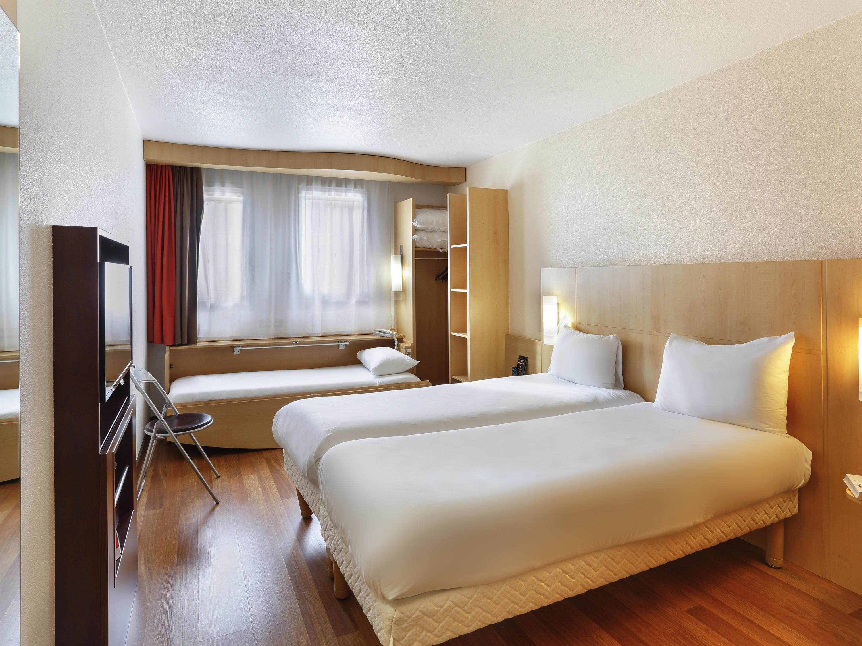 Een hotelkamer met twee bedden, een televisie, een kledingkast, een spiegel en een raam met gordijnen.
