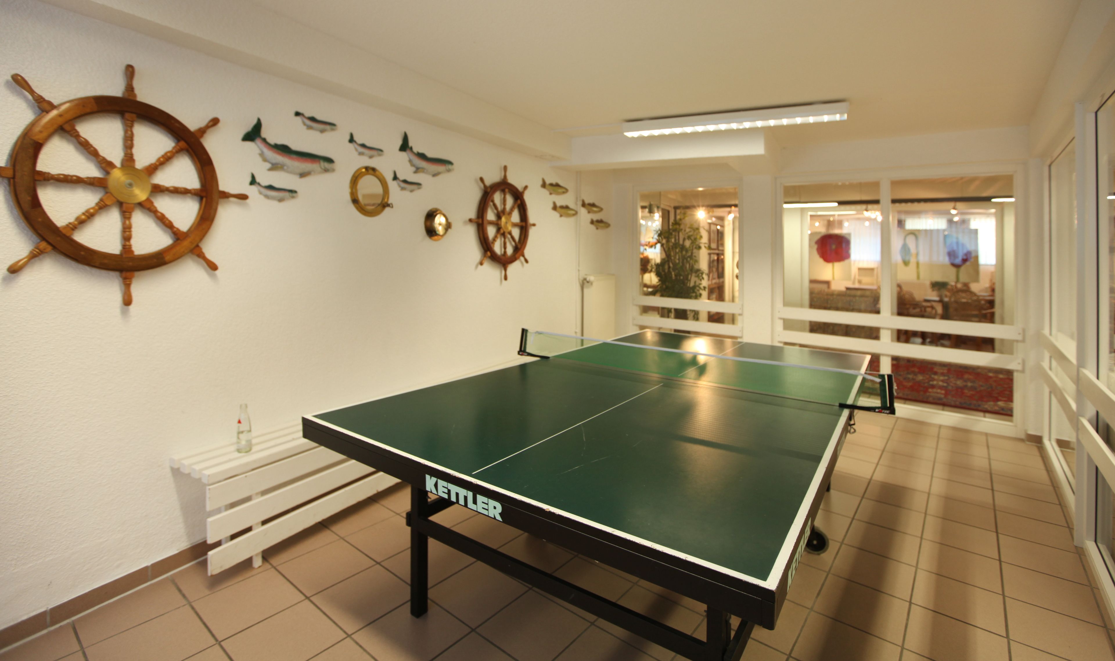 Una mesa de ping pong con la etiqueta 'Kettler', una rueda de barco y varias decoraciones de peces en la pared.