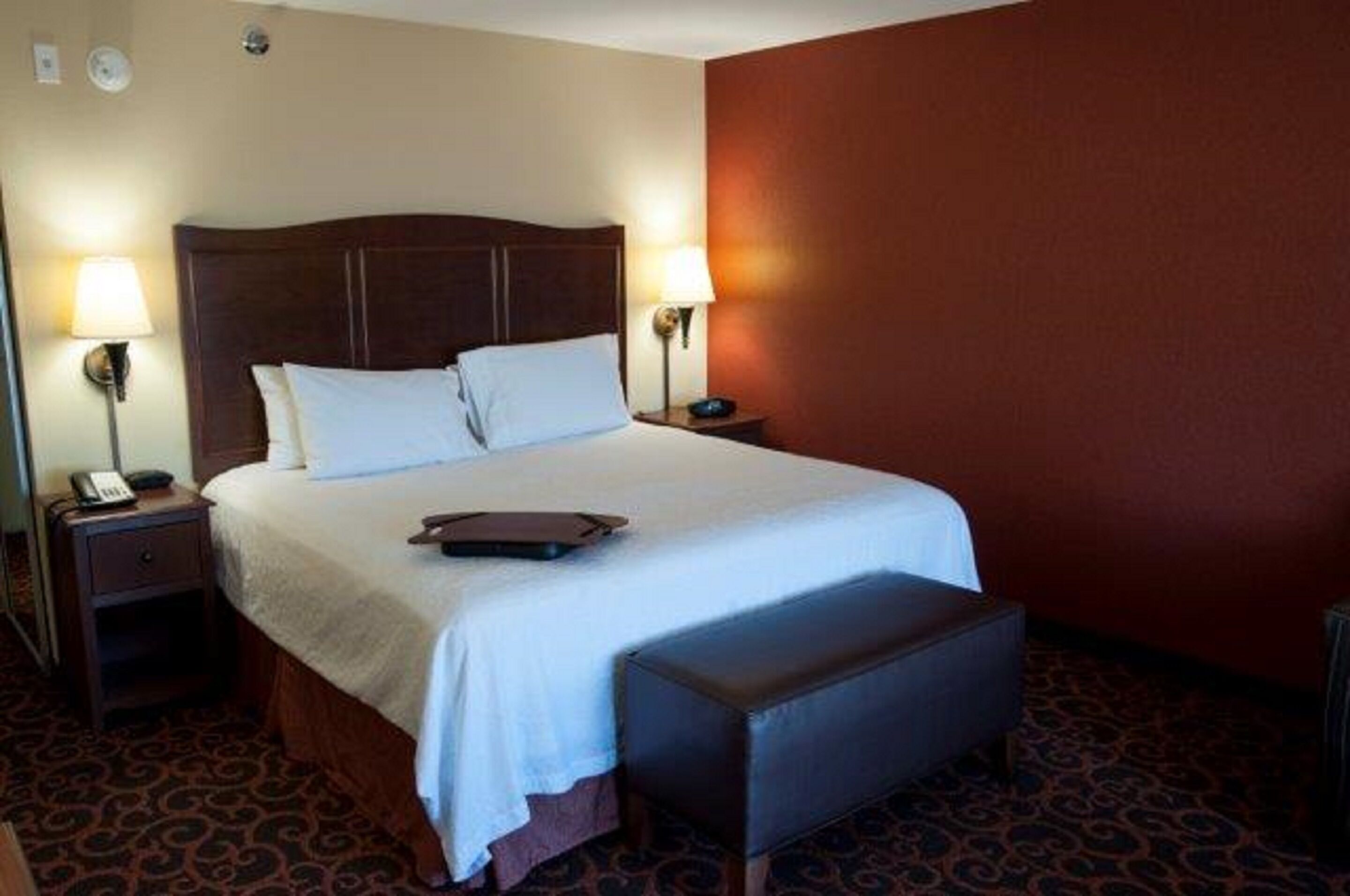A hotel room with a large bed, two bedside tables, a nightstand, a bench, and a patterned carpet.