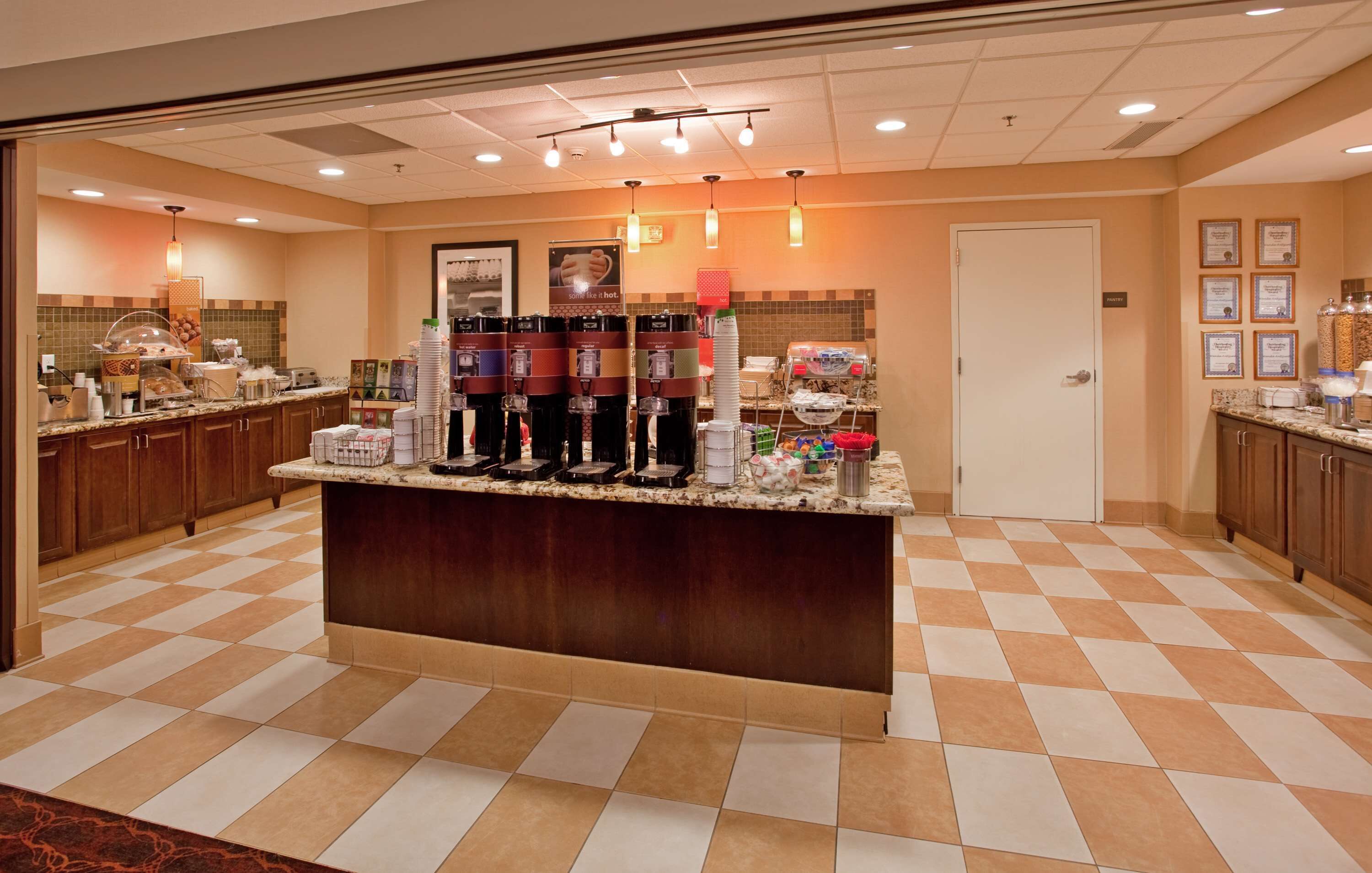 A hotel lobby with a checkered floor, a central counter with beverage dispensers, and a door in the background.