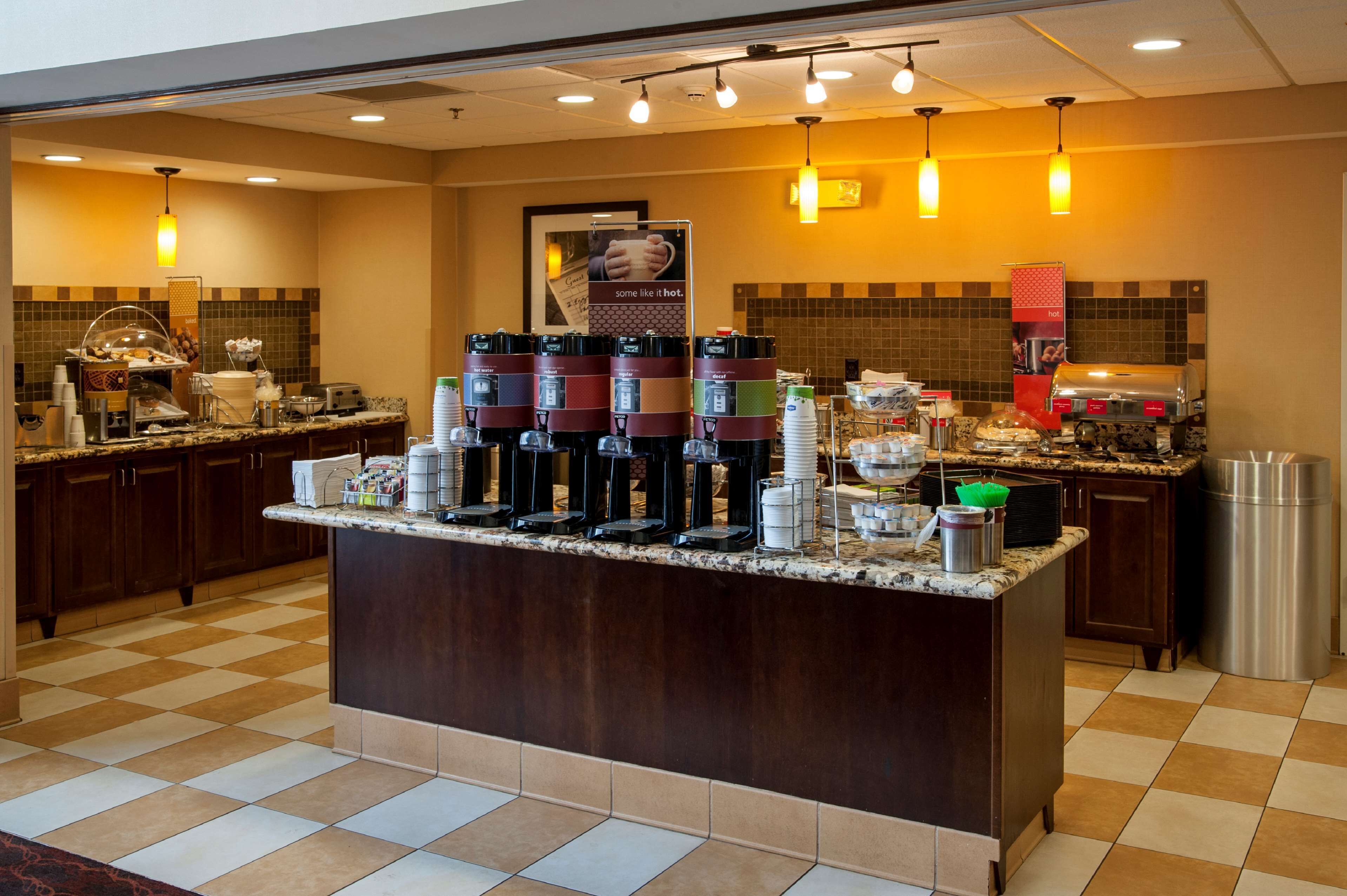 A hotel breakfast area with a self-serve coffee station, buffet, and a large poster on the wall.