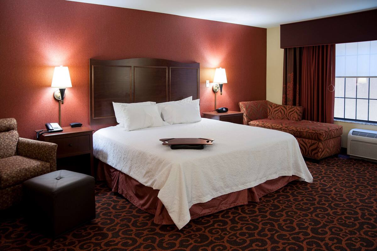 A hotel room with a large bed, two bedside lamps, a chair, an ottoman, and a window with curtains.