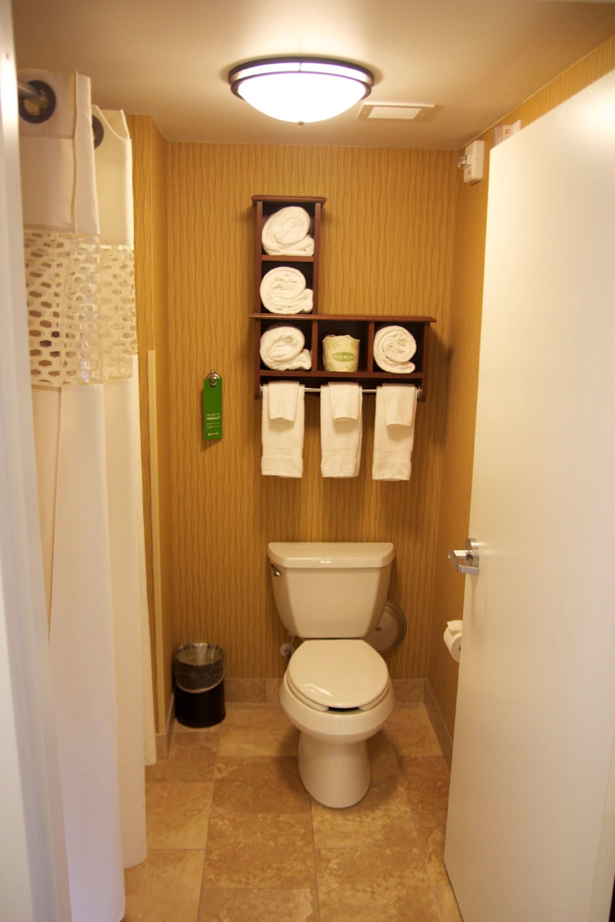 Een kleine badkamer met een toilet, een plank met opgevouwen handdoeken en een vuilnisbak.