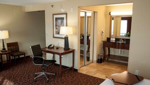 A hotel room with a bed, a desk with a chair, a telephone, a lamp, a mirror, and a bathroom visible through a glass door.