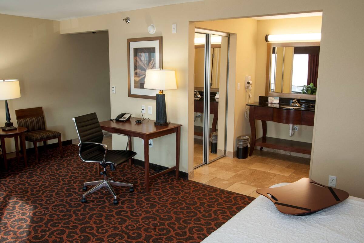A hotel room with a bed, a desk with a chair, a telephone, a lamp, a mirror, and a bathroom visible through a glass door.